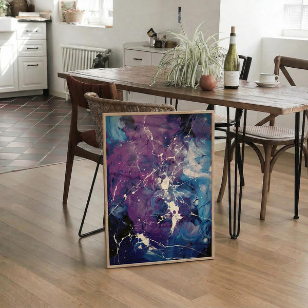 May include: Abstract artwork with purple and blue fluid paint splashes and white marble-like veining, displayed in a light wood frame leaning against a rustic wooden dining table. The poster shows flowing, organic shapes with deep violet and cobalt blue tones blending together. Kitchen setting with woven chairs, plant, and wine bottle visible in background on light wood floors.