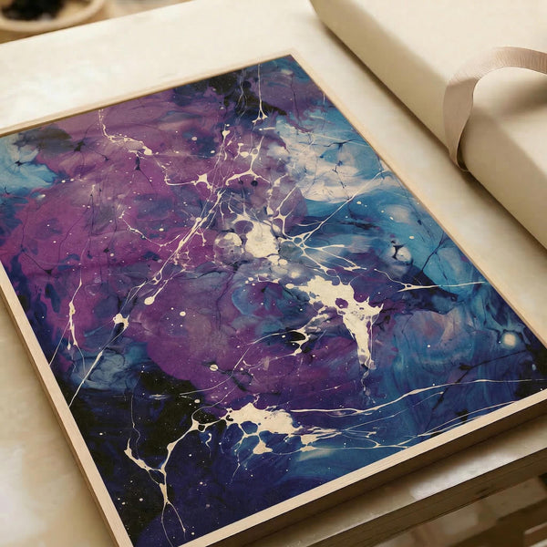 Abstract fluid art print with flowing purple, blue, and white paint creating organic shapes and delicate crackling lines throughout. The artwork features marbled textures with splashes and drips, displayed flat on white paper with a roll of paper visible in the background on a light wooden surface.