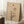 May include: Framed minimalist artwork featuring a bare black tree silhouette against beige textured background with rust-colored sun, large tan circle, and subtle geometric lines. Light wood frame leaning against white wall near white coffee cup on wooden block and cream textured throw blanket.