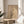 May include: Framed abstract minimalist artwork displayed on a round wooden side table beside a beige upholstered chair. The poster features a silhouetted bare tree against neutral beige tones with geometric shapes including circles and a small red sun element. Natural sunlight streams through white curtains creating soft shadows on the wall. A plant leaf is partially visible in the corner, adding to the serene modern interior setting.
