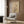 May include: Beige armchair with geometric pillow in modern living room setting. Abstract minimalist poster on wood-paneled wall features bare tree silhouette with geometric shapes - circles and rectangles in neutral tones with small red accent dot. Gold floor lamp, dark round side table with open book, and sheer curtains complete the contemporary interior scene.