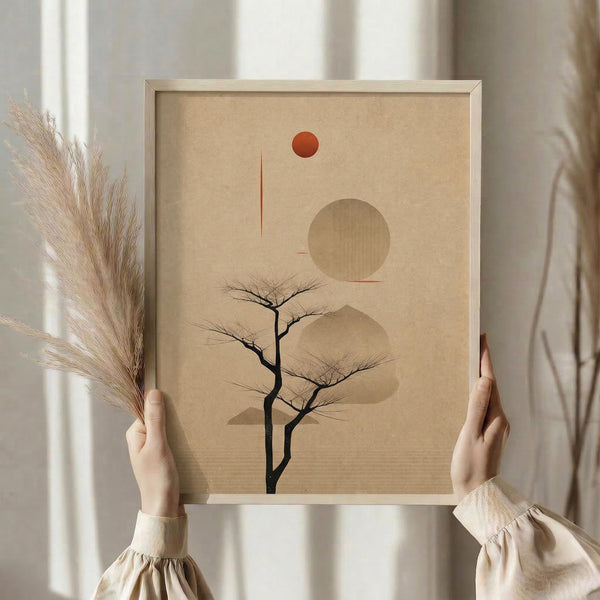 May include: Hands holding a framed minimalist artwork featuring a bare black tree silhouette against beige background with geometric shapes - red circle sun, large tan circle, and curved mountain forms. Light wood frame, soft natural lighting, dried pampas grass visible to left, cream curtains in background.