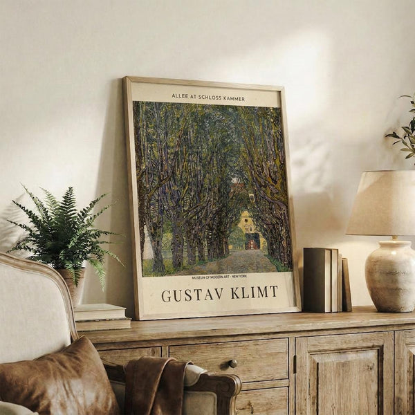 May include: Framed art print featuring a tree-lined path leading to a small building, displayed on a rustic wooden console table alongside potted fern, ceramic vase, books, and brown leather chair in a neutral-toned living room setting.
