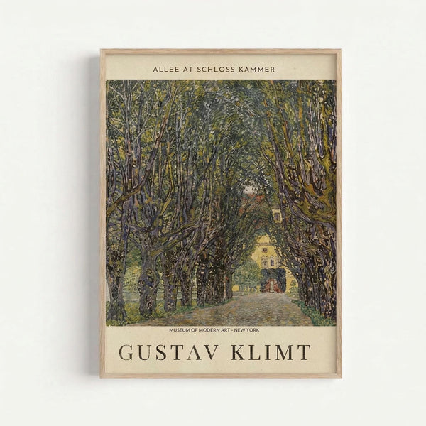 May include: Framed art print mounted on white wall showing tree-lined pathway leading to yellow building. Dense canopy of green and brown trees creates natural tunnel effect over cobblestone path. Vintage-style poster with cream matting displays "GUSTAV KLIMT" and "MUSEUM OF MODERN ART - NEW YORK" text. Light wood frame complements earth-toned artwork with impressionistic brushwork and rich forest greens.