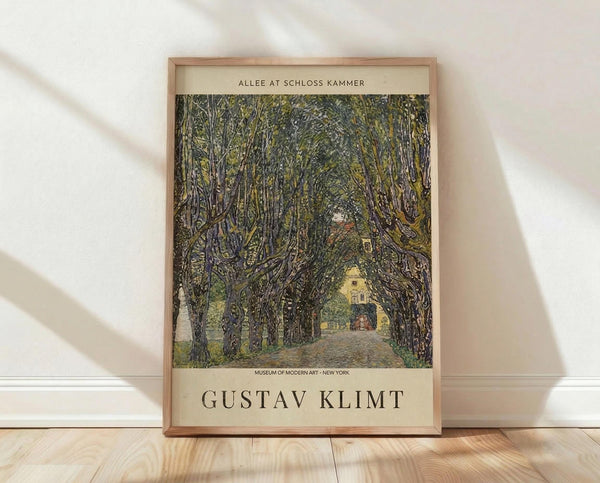 May include: Framed vintage art poster featuring a tree-lined avenue leading to a yellow building, displayed in thin wooden frame against white textured wall on light wood flooring with natural shadows. The artwork shows detailed green foliage creating a tunnel effect over a dirt path, with museum attribution text above and artist name Gustav Klimt below the image.