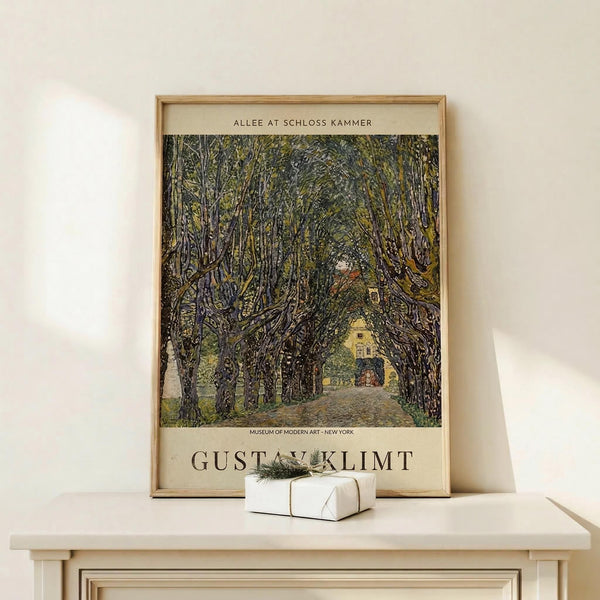 May include: Framed art print featuring a tree-lined avenue leading to a yellow building, painted in an impressionistic style with thick brushstrokes in greens, browns, and muted tones. The poster shows "Gustav Klimt" text and "Museum of Modern Art - New York" attribution. Displayed on a light wood surface with a small white wrapped gift beside it, leaning against a cream-colored wall in natural lighting.