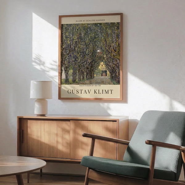 May include: Framed Gustav Klimt museum poster showing a tree-lined pathway leading to a yellow building, displayed above a wooden sideboard with white ceramic lamp. Mid-century modern green upholstered chair with wooden arms sits nearby. Warm sunlight creates shadows across white wall and wooden floor in minimalist living room setting.