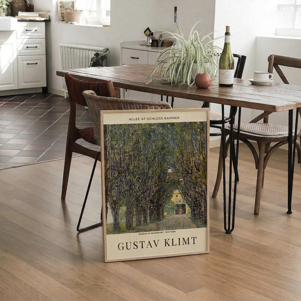 May include: A framed art print leaning against a wooden dining table in a modern kitchen. The poster shows a tree-lined pathway leading to a yellow building, painted in earthy greens and browns. The print features Gustav Klimt's name and Museum of Modern Art attribution at the bottom. The scene includes wicker dining chairs, a spider plant, wine bottle, and white kitchen cabinets in the background on light wooden floors.