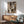 May include: Art deco style portrait poster featuring geometric woman's face in earth tones - browns, greens, and oranges - displayed in thin wooden frame above mid-century modern credenza. Scene shows teal upholstered armchair with wood arms, white ceramic table lamp, round coffee table, and natural sunlight casting shadows on white wall.