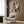 May include: Stylized Art Deco woman portrait poster in warm earth tones - browns, oranges, and greens - featuring geometric cubist-style face with angular features. Artwork shown framed in light wood frame, displayed on wood-paneled wall above cream velvet armchair with patterned pillow. Modern brass floor lamp and side table with open book complete the sophisticated neutral-toned room setting.