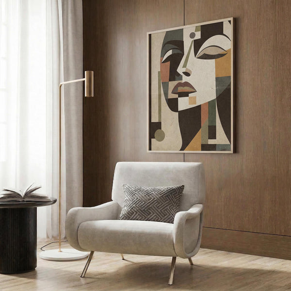May include: Stylized Art Deco woman portrait poster in warm earth tones - browns, oranges, and greens - featuring geometric cubist-style face with angular features. Artwork shown framed in light wood frame, displayed on wood-paneled wall above cream velvet armchair with patterned pillow. Modern brass floor lamp and side table with open book complete the sophisticated neutral-toned room setting.