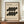 May include: Framed Bauhaus-inspired poster with geometric black kidney-shaped forms arranged in two rows on cream background, dated 1919, displayed on wooden credenza next to white ceramic vase and stacked books in neutral-toned room with white paneled walls.