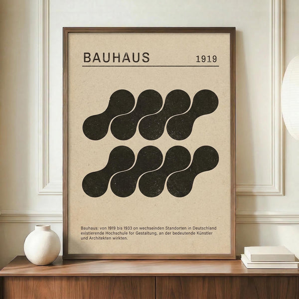 May include: Framed Bauhaus-inspired poster with geometric black kidney-shaped forms arranged in two rows on cream background, dated 1919, displayed on wooden credenza next to white ceramic vase and stacked books in neutral-toned room with white paneled walls.