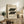 May include: Framed Bauhaus poster with cream background and black geometric shapes arranged in two rows, dated 1919, displayed on rustic wooden sideboard. Room features neutral tones, fern plant, pottery vases, books, and beige upholstered chair with brown leather pillow. Natural lighting creates warm, minimalist interior styling with earthy textures.
