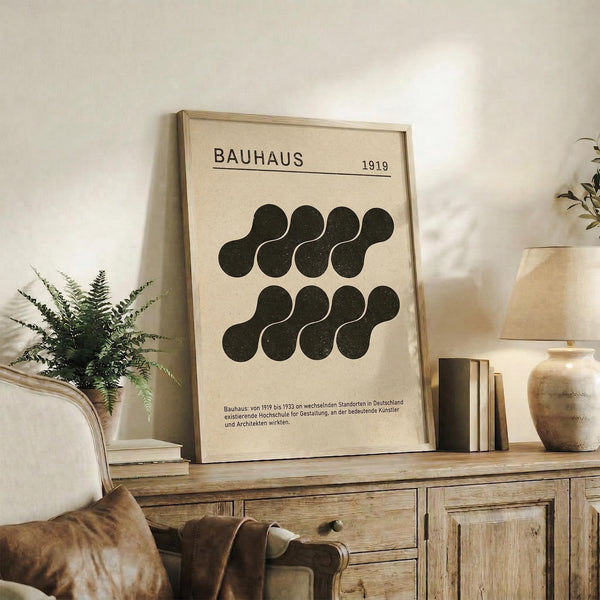 May include: Framed Bauhaus poster with cream background and black geometric shapes arranged in two rows, dated 1919, displayed on rustic wooden sideboard. Room features neutral tones, fern plant, pottery vases, books, and beige upholstered chair with brown leather pillow. Natural lighting creates warm, minimalist interior styling with earthy textures.