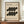 May include: Framed Bauhaus-inspired poster with cream background featuring black organic curved shapes arranged in two rows, dated 1919, with German text below. Wood frame sits on wooden console beside white ceramic vase and stacked books against cream paneled wall.