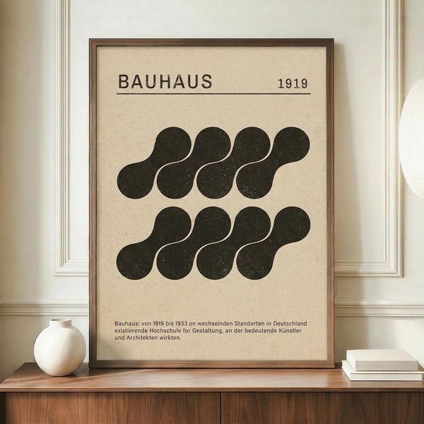 May include: Framed Bauhaus-inspired poster with cream background featuring black organic curved shapes arranged in two rows, dated 1919, with German text below. Wood frame sits on wooden console beside white ceramic vase and stacked books against cream paneled wall.