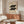 May include: Vintage-style Bauhaus poster dated 1919 featuring black geometric circular shapes arranged in two rows on cream background, displayed above mid-century modern walnut credenza with brass handles. Room setting shows white upholstered chair, dome table lamp, oval glass coffee table, and warm neutral walls with natural lighting creating soft shadows.