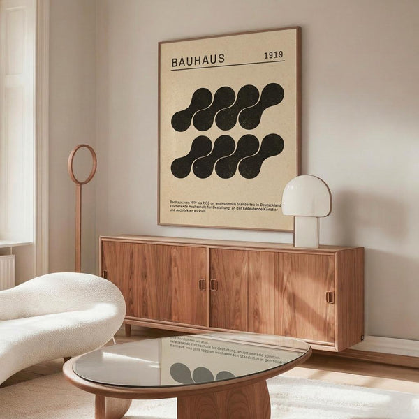 May include: Vintage-style Bauhaus poster dated 1919 featuring black geometric circular shapes arranged in two rows on cream background, displayed above mid-century modern walnut credenza with brass handles. Room setting shows white upholstered chair, dome table lamp, oval glass coffee table, and warm neutral walls with natural lighting creating soft shadows.
