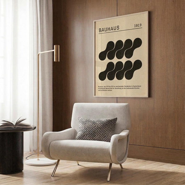 May include: Modern living room with beige armchair and patterned pillow, featuring vintage Bauhaus 1919 poster on wood-paneled wall. Poster displays black geometric circles arranged in rows on cream background with German text. Scene includes brass floor lamp, dark side table with open book, and white curtains. Warm wood tones throughout.