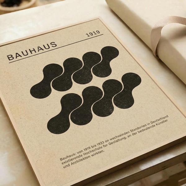 Minimalist cream-colored poster featuring black organic blob shapes arranged in a circular pattern. The design shows "BAUHAUS" and "1919" text at the top with German text below describing the influential design school. The poster lies flat on white paper with rolled paper tubes visible in the background, showing the clean modernist aesthetic typical of Bauhaus design principles.