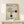May include: Framed Bauhaus-inspired poster with geometric design featuring concentric circles, parallel lines, and two black dots on cream background, displayed on light wood console table with small white wrapped gift, against neutral wall with natural lighting