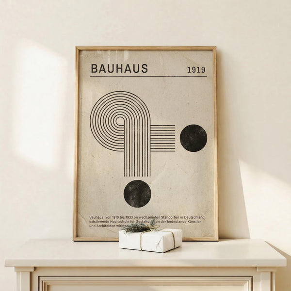 May include: Framed Bauhaus-inspired poster with geometric design featuring concentric circles, parallel lines, and two black dots on cream background, displayed on light wood console table with small white wrapped gift, against neutral wall with natural lighting