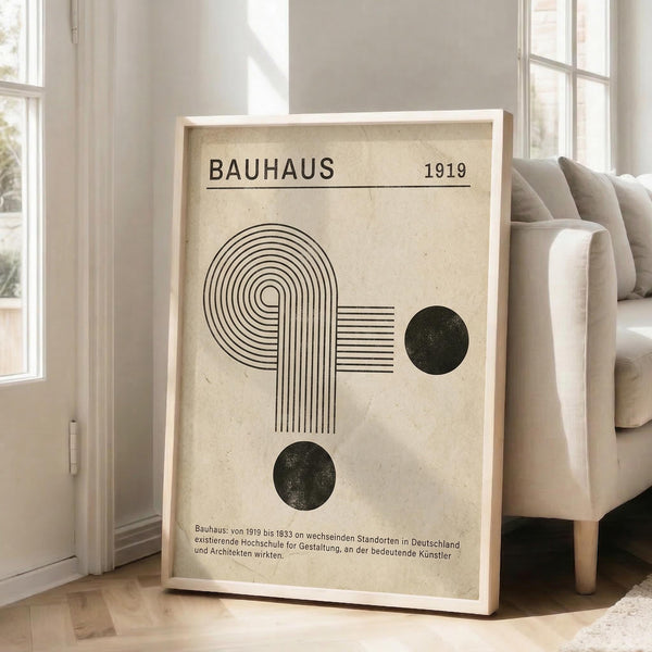 May include: Framed Bauhaus-inspired poster with geometric design featuring curved lines forming an abstract shape, two dark circles, and "BAUHAUS 1919" text on cream background with German text below. Light wood frame leaning against white wall next to beige sofa in bright, minimalist living room with large windows.