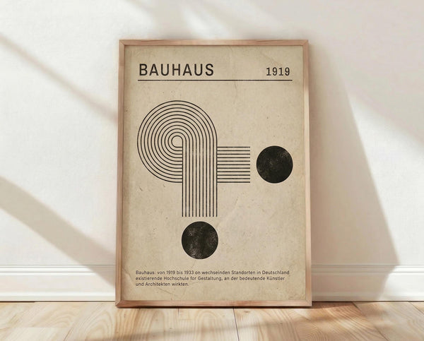 May include: Framed Bauhaus-inspired poster with geometric design featuring curved lines forming an abstract shape, two black circles, and "BAUHAUS 1919" text on cream background with German text below, displayed in light wood frame leaning against white wall on wooden floor with soft natural lighting