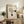 May include: Framed beige poster with "BAUHAUS 1919" text and geometric black line design featuring curved and straight elements with two black circles, displayed on rustic wooden sideboard alongside potted fern, ceramic vases, and books in warm neutral living room setting with leather furniture