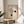 May include: Framed Bauhaus-inspired minimalist poster with geometric spiral design and black circles on cream background, displayed on round wooden table beside white upholstered chair and potted plant in bright, sunlit room with neutral walls and flowing curtains.