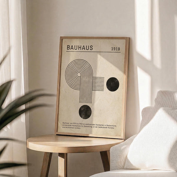May include: Framed Bauhaus-inspired minimalist poster with geometric spiral design and black circles on cream background, displayed on round wooden table beside white upholstered chair and potted plant in bright, sunlit room with neutral walls and flowing curtains.