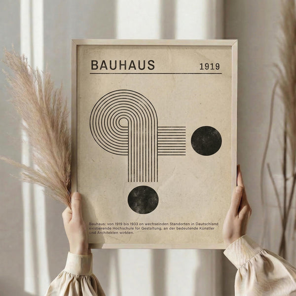 May include: Person in beige clothing holding framed Bauhaus poster with geometric design featuring curved lines forming abstract shapes and two black circles on cream background, dated 1919, with dried pampas grass visible beside them against neutral curtained backdrop