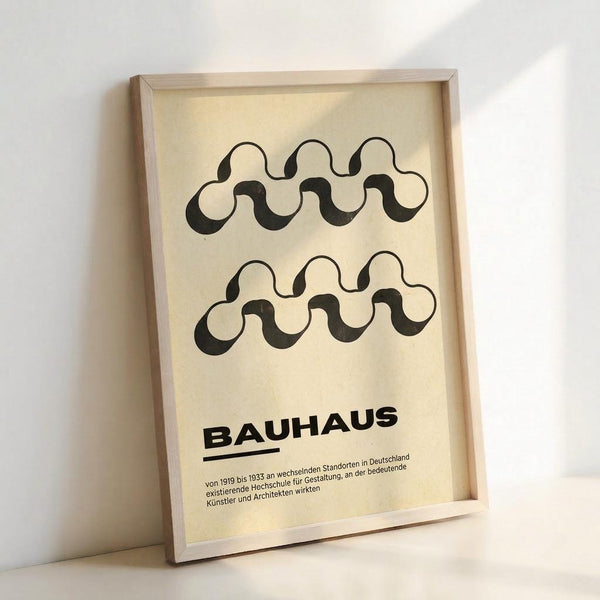 May include: Framed poster with cream background featuring two black wavy serpentine shapes arranged horizontally. Bold "BAUHAUS" text below with German descriptive text underneath. Light wooden frame leaning against white wall with natural lighting creating soft shadows.
