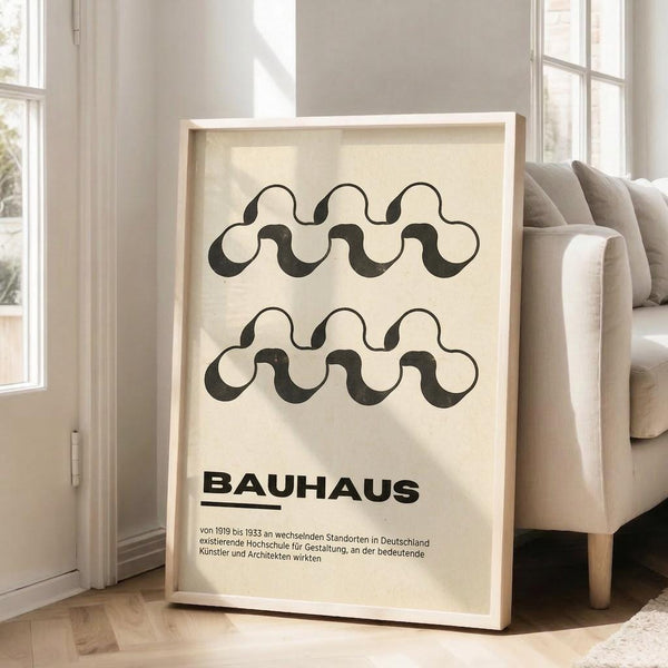 May include: Framed Bauhaus-style poster with black wavy interlocking pattern on cream background, displayed on wooden floor next to beige armchair in bright living room with white walls and large windows. The minimalist geometric design features two rows of flowing wave forms above bold text in German describing the art school's history from 1919-1933.