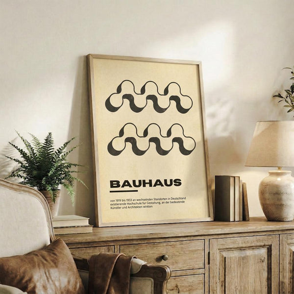 May include: Framed poster with cream background displaying two rows of black wavy interlocking chain patterns above bold "BAUHAUS" text and German description. Natural wood frame sits on rustic wooden sideboard alongside fern plant, ceramic vase, stacked books, and partial view of tan leather armchair in warm neutral living room setting.