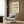 May include: Cream-colored poster with black wavy abstract design and "BAUHAUS" text displayed on wood-paneled wall above beige velvet armchair with geometric patterned pillow, modern brass floor lamp, and black side table with open book in contemporary living room setting with natural lighting from window.