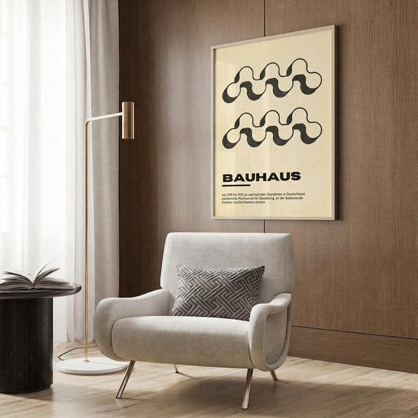 May include: Cream-colored poster with black wavy abstract design and "BAUHAUS" text displayed on wood-paneled wall above beige velvet armchair with geometric patterned pillow, modern brass floor lamp, and black side table with open book in contemporary living room setting with natural lighting from window.