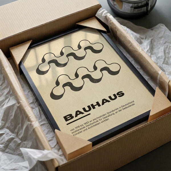 Framed poster with cream background featuring abstract wavy black lines in two horizontal rows above bold "BAUHAUS" text with German description below. Black frame sits in cardboard shipping box with white tissue paper padding on wooden surface next to wrapped tube.