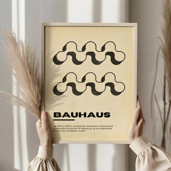 May include: Person in beige clothing holding framed poster with cream background featuring two rows of black wavy interlocking patterns above "BAUHAUS" text and German description. Silver frame, pampas grass visible to left, soft natural lighting from white curtained window.