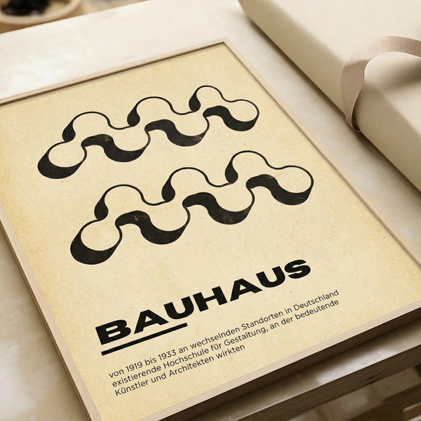 May include: Cream-colored poster with black wavy line pattern displayed in white frame on neutral surface. Design features flowing serpentine shapes above bold "BAUHAUS" text with German descriptive text below. Minimalist geometric artwork shown alongside white ribbon, suggesting gift presentation or styling context.