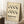 May include: Framed poster with cream background featuring black wavy interlocking chain-like patterns and "BAUHAUS" text with German description below. Light wood frame leans against white paneled wall next to beige textured chair, with coffee cup on wooden side table and white fuzzy rug beneath.