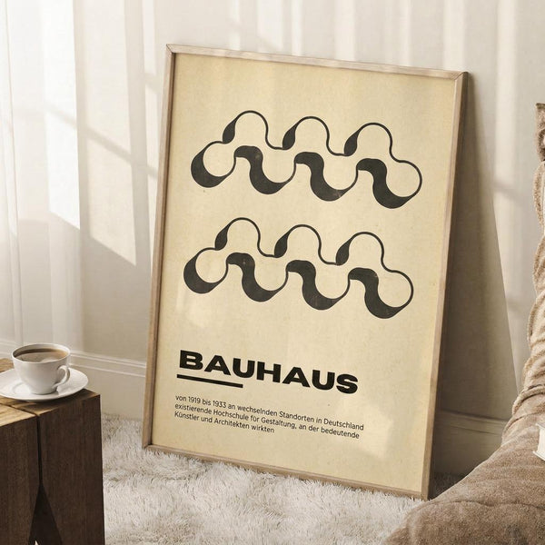 May include: Framed poster with cream background featuring black wavy interlocking chain-like patterns and "BAUHAUS" text with German description below. Light wood frame leans against white paneled wall next to beige textured chair, with coffee cup on wooden side table and white fuzzy rug beneath.