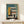May include: Framed Bauhaus-style architectural poster showing geometric building design in teal, mustard yellow, coral, and cream colors with "02" marking, displayed on light wood surface next to small wrapped gift with twine and greenery sprig against white wall