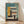 May include: Framed modernist architectural poster featuring geometric building illustration in teal, yellow, orange and cream colors with "BAUHAUS" text and "02" numbering. Wood frame leaning against white wall on light wooden floor with natural window shadows.