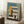 May include: Framed Bauhaus-style architectural poster with geometric building design in mustard yellow, coral, teal and cream colors, displayed in light wood frame leaning against white wall corner. Coffee cup on wooden block and textured throw blanket visible in cozy interior setting with natural window light creating shadows.