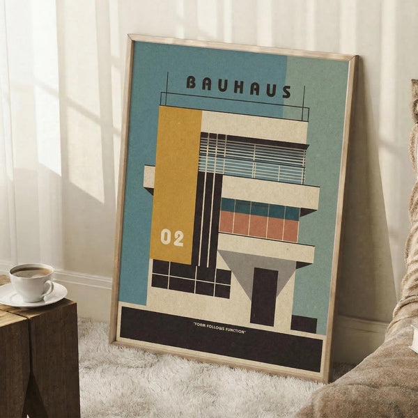 May include: Framed Bauhaus-style architectural poster with geometric building design in mustard yellow, coral, teal and cream colors, displayed in light wood frame leaning against white wall corner. Coffee cup on wooden block and textured throw blanket visible in cozy interior setting with natural window light creating shadows.