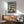 May include: Modernist architectural poster with teal background showing geometric building design in yellow, white, and black. Features "Bauhaus" text and "02" numbering. Displayed in thin copper frame above wooden sideboard with white ceramic lamp. Mid-century modern sage green armchair with wooden arms in foreground. Bright natural light creates shadows across white wall.