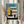 May include: Person holding framed architectural poster featuring modernist building design in geometric style with yellow, teal, coral and cream color blocks, horizontal window details, and "Form Follows Function" text at bottom. Natural lighting, pampas grass decoration visible, light wood frame against neutral interior background.
