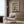 May include: Framed Bauhaus-style architectural poster featuring a modernist building in teal, yellow, orange and cream geometric shapes, displayed above a beige upholstered armchair with patterned pillow. Room has wood paneled walls, brass floor lamp, and dark round side table with open book. Poster shows clean lines and bold color blocks typical of Bauhaus design movement.